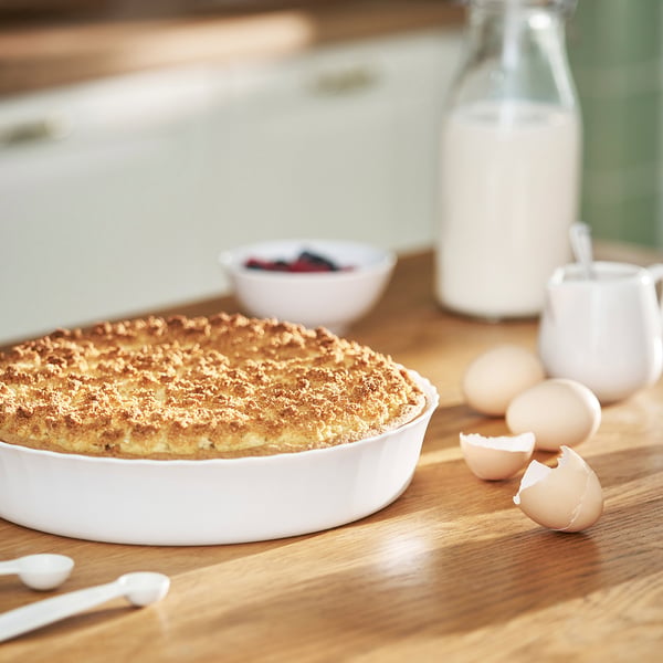 A baked dish in a white ceramic pie plate sits on a wooden table, surrounded by ingredients including eggs, a jar of milk, and a bowl of berries.