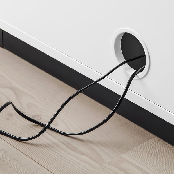 Black power cable exiting circular hole in white cabinet, lying on wooden floor.