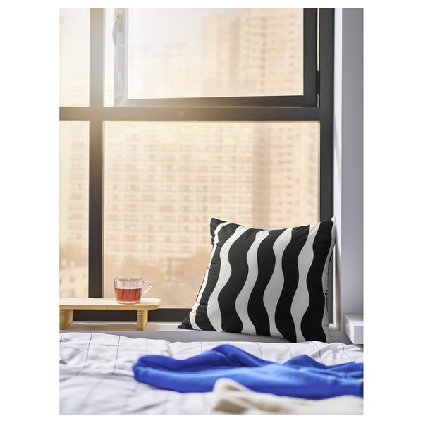 Bed with a black and white striped cushion and a blue blanket; a tea cup on a wooden table beside a window.