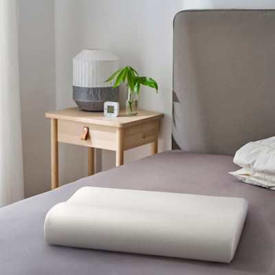 Modern bedroom featuring a white GRÖNPIL pillow on a grey bed next to a wooden bedside table with a white lamp and green plant.
