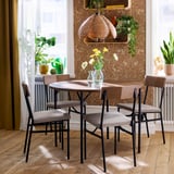 Modern dining set with round table and four chairs. Light wood top, black metal base. Upholstered seats, wooden backs. Vase with flowers.