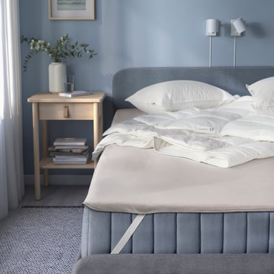 Blue bedroom with waterproof mattress protector under white sheets, beside wooden bedside table.