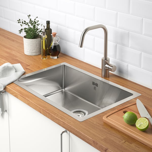 Modern stainless steel sink with sleek tap on wooden countertop. Limes and knife on cutting board; bottles and plant nearby.