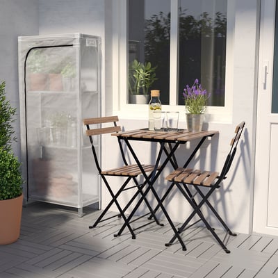 Small outdoor patio set with wooden table and two chairs, near a white wall with a window and a potted plant.