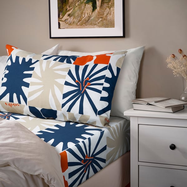 Bed with floral HYLSNEJLIKA covers, envelope pillowcases, small drawer beside.