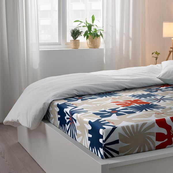Bed with colourful floral-patterned bedding, white pillows, and plants on the windowsill.
