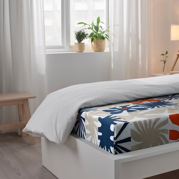 Bright bedroom with white bed, colourful floral bedspread, two plants by the window.