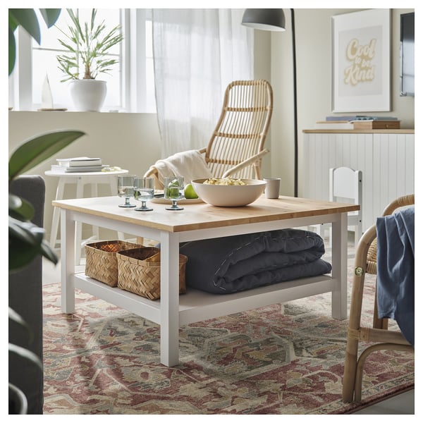 Modern living room with wooden table, wine glasses, blanket, wicker chairs, pastel rug, plant, & cool to be kind poster.