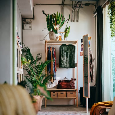 Green hanging storage bag on wooden rack, among coats in plant-decorated room.