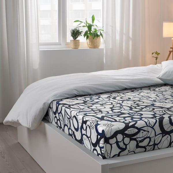 Bed with white comforter, patterned sheet, two potted plants near window, sheer curtains.