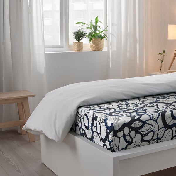 Bedroom with white bed, patterned sheets, white pillows, wooden bench, and plants on windowsill.