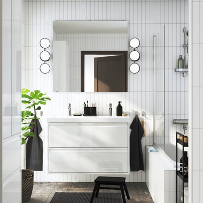 Modern bathroom: large mirror with lights, white vanity, black towels, glass shower, plant.