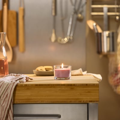 Small pink scented candle in a glass cup on a wooden shelf with cooking utensils.