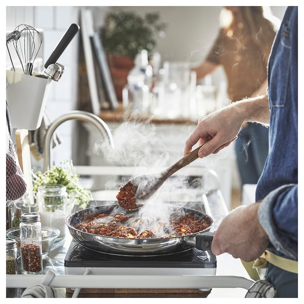 A person cooks food in a frying pan on a stove in a kitchen. There is steam rising from the pan and various kitchen utensils and ingredients around.