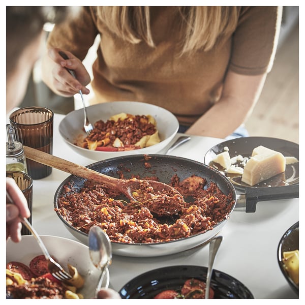 A person serves cooked food from a MIDDAGSMAT pan on a table with plates and drinks. The pan is brown with a wooden handle.