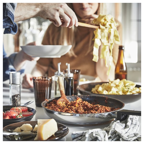 Family cooking meal, person serving stew, dishes, glasses, bottle, cheese, tomato, herbs on table.