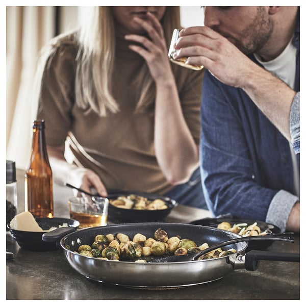 Two people dining, enjoying food and drinks. In focus is a black frying pan with cooked brussels sprouts.
