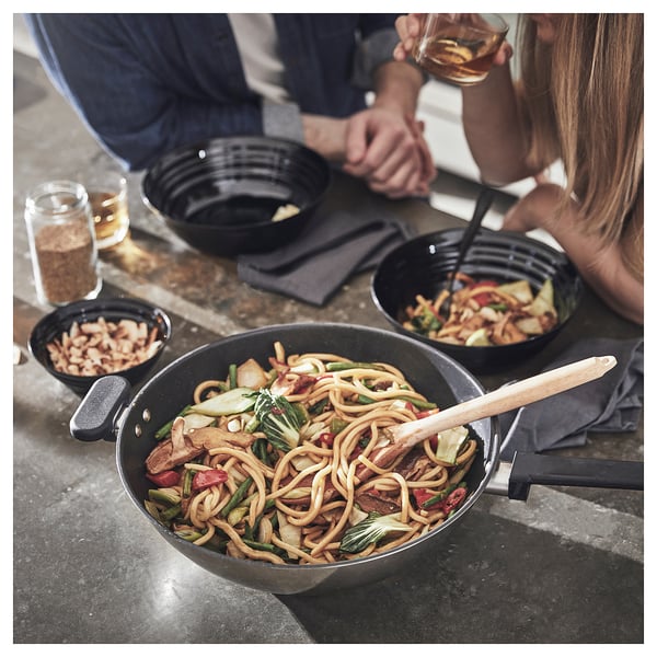 Two people eating noodles from black bowls at a table with drinks, using chopsticks. A black skillet with mixed noodles is nearby.