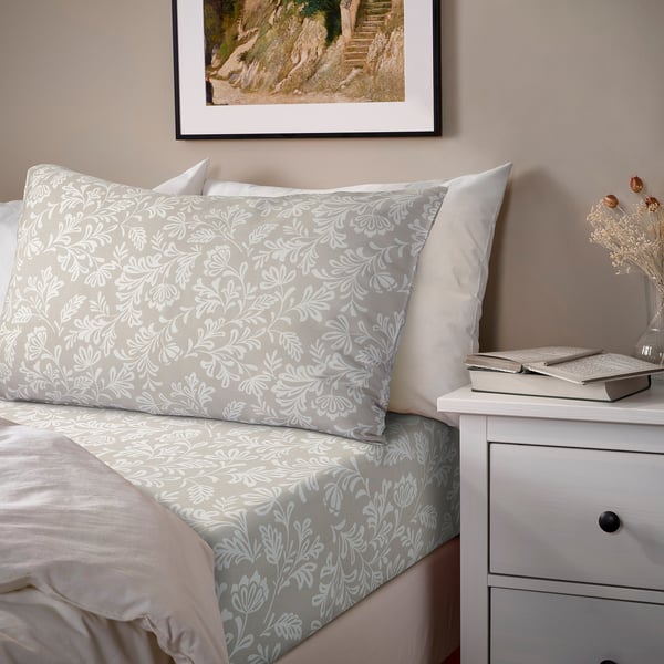 Made bed with grey floral sheets & pillowcases, small white dresser, book & vase, framed pic above.