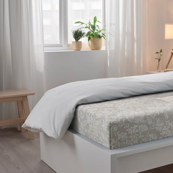 Bedroom with a white bed, patterned bedsheet, wooden bench, and two potted plants on the windowsill. Sheer white curtains let in soft light.