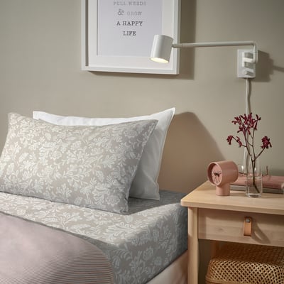 Cosy bedroom with grey floral bedding, bedside table, pink clock, and red flowers.