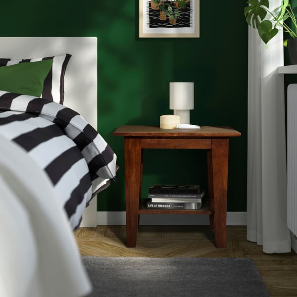 NORRVÅGA bedside table in bedroom with books, lamp, candle, & striped bedding against green wall.