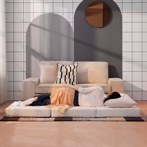 Person relaxing on a beige modular sofa with cushions, which can transform into a guest bed. The room has an arched backdrop with white tiled walls.