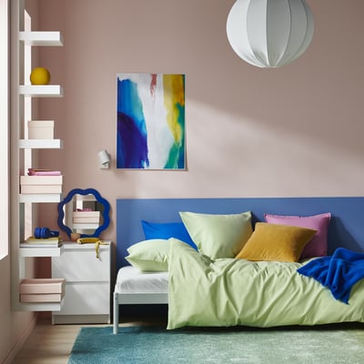 Modern bedroom with PILTANDVINGE bedding, lime green covers, blue pillows & blanket. Side table with books and mirror. Colourful wall art.
