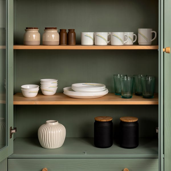 Green kitchen cabinet with three shelves: jars, mugs, bowls, plates, glasses, vase, canisters.