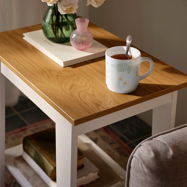 Small white SKRÄNTÄRNA mug with blue floral design on a wooden table, next to a green vase and pink glass, beside a book and chair.