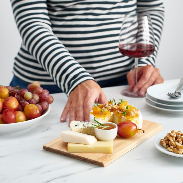 Person with cheese platter on bamboo board, red wine, fruit, and nuts.