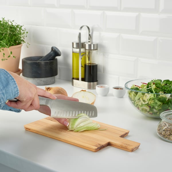 Person slicing onion on bamboo cutting board next to mixed salad bowl, herbs, and oils.