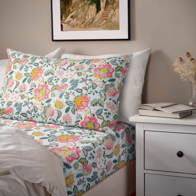 Floral bed with SOCKERBAGARE sheet set. White base, matching pillowcases, small bedside table with laptop, framed picture.