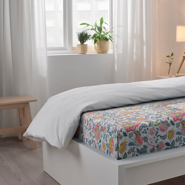 Bed with white sheet and floral pattern, two plants on windowsill, light curtains, small wooden stool by bed.