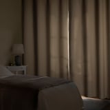 Dim bedroom with grey bed, small white bedside table, and floor-to-ceiling curtains.