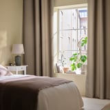 Bedroom with large window, beige curtains block light, modern lamp on bedside table, green plant on windowsill.