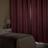 Bedroom with dark red curtains blocking light, a white bedside table, and a lamp.