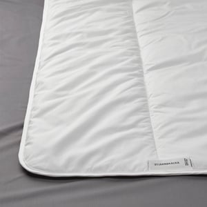 Close-up of a white, plush comforter with visible stitching, neatly spread out on bed. Brand label visible at bottom.