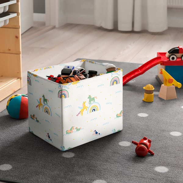 Decorative toy box with unicorn design on floor, surrounded by children’s toys and wooden furniture.