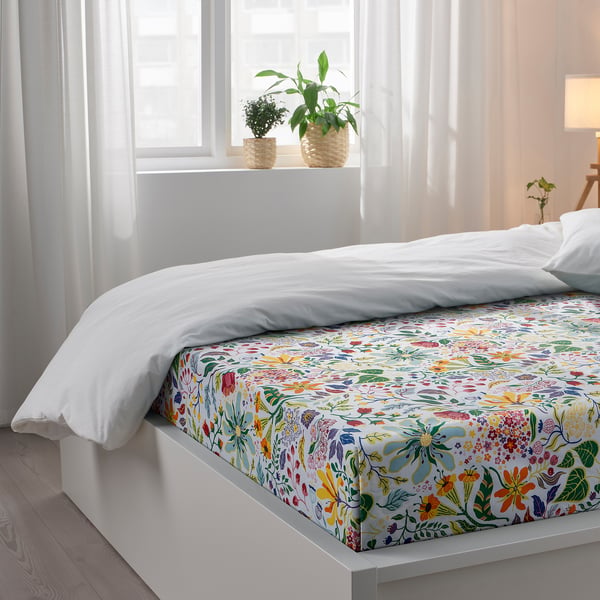 A cosy bedroom with a white bed featuring a floral-printed flat sheet and pillowcases made of 100% cotton. Plants rest on the windowsill above.