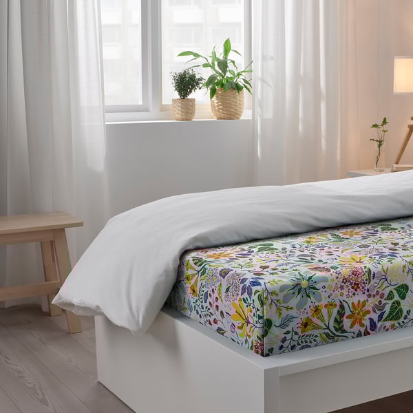 Bright, airy bedroom with a white bed featuring colourful floral bedding, two potted plants on the windowsill, and sheer white curtains.