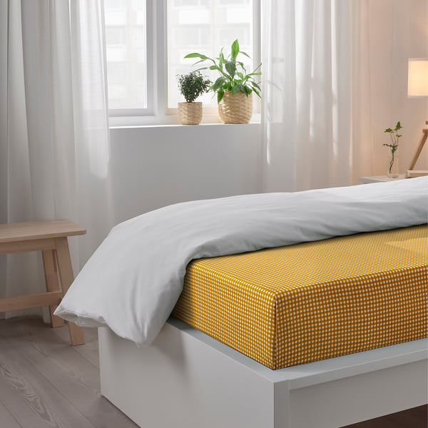 Bright bedroom with a bed covered in a yellow-checkered sheet set by TETSTJERNA, featuring white pillows with a wooden bench and a window with plants.