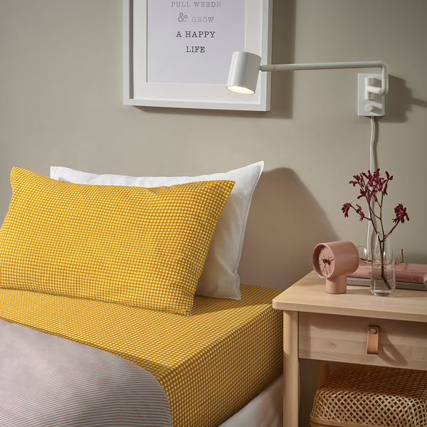 A cosy bedroom corner featuring a bed with yellow chequered TETSTJERNA sheets, a modern wall lamp, and a bedside table with a pink clock and vase.