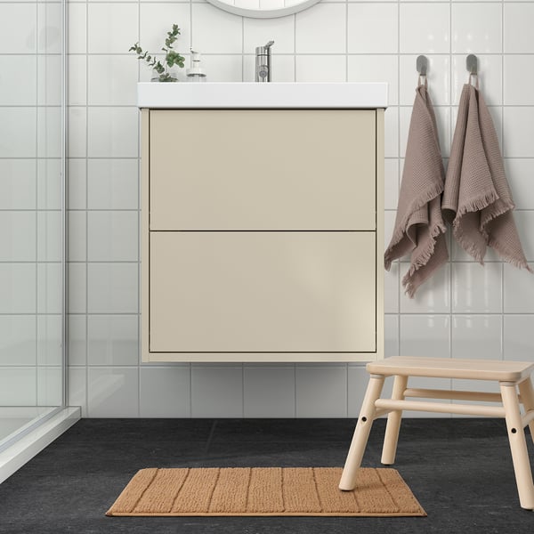 Bathroom scene: beige TIDNINGSBUD mat, light-colored wooden stool, beige cabinet, towels, glass door. Mat is small, beneath stool.