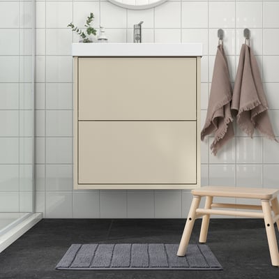 Modern bathroom with large beige cabinet, light grey tiled walls, wooden bench, grey bath mat, and beige towels hanging.