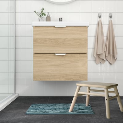 Modern bathroom with light wood cabinet, white sink, beige tiles, blue mat, small stool, & hanging towels.