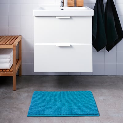 White bathroom cabinet with sink, blue bath mat on floor, wooden bench with towels, grey tiled walls.
