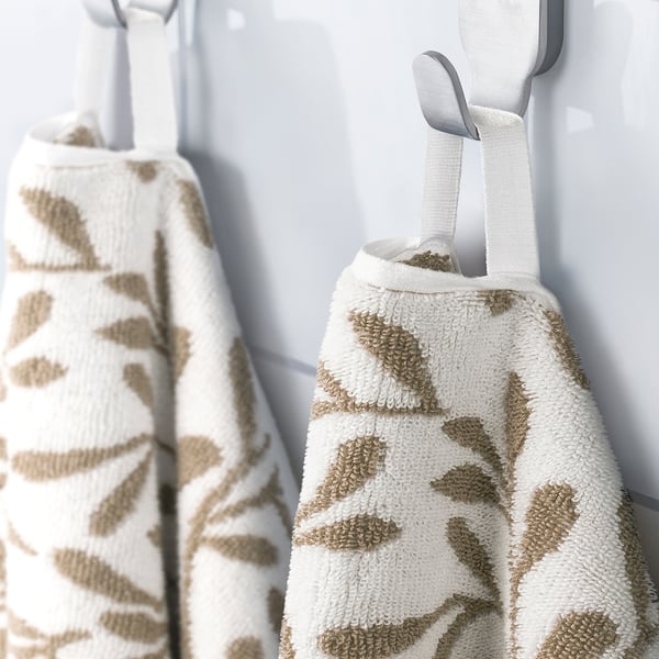 Two white towels with beige leaf patterns hang on hooks. One in foreground, showing loop for hanging.