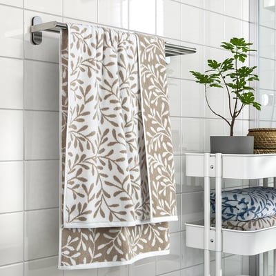 White towel with brown leaf pattern hangs on silver rail, next to green plant on white stand.