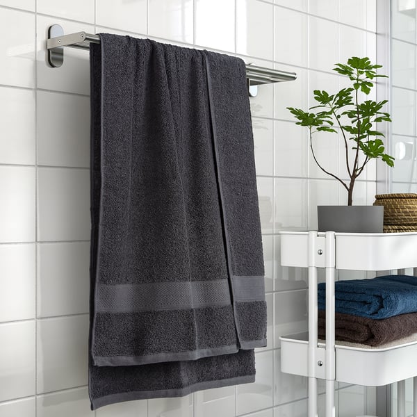Dark grey VITBACKEN bath towel hanging on silver rail, larger than folded towels on white shelf with plant.